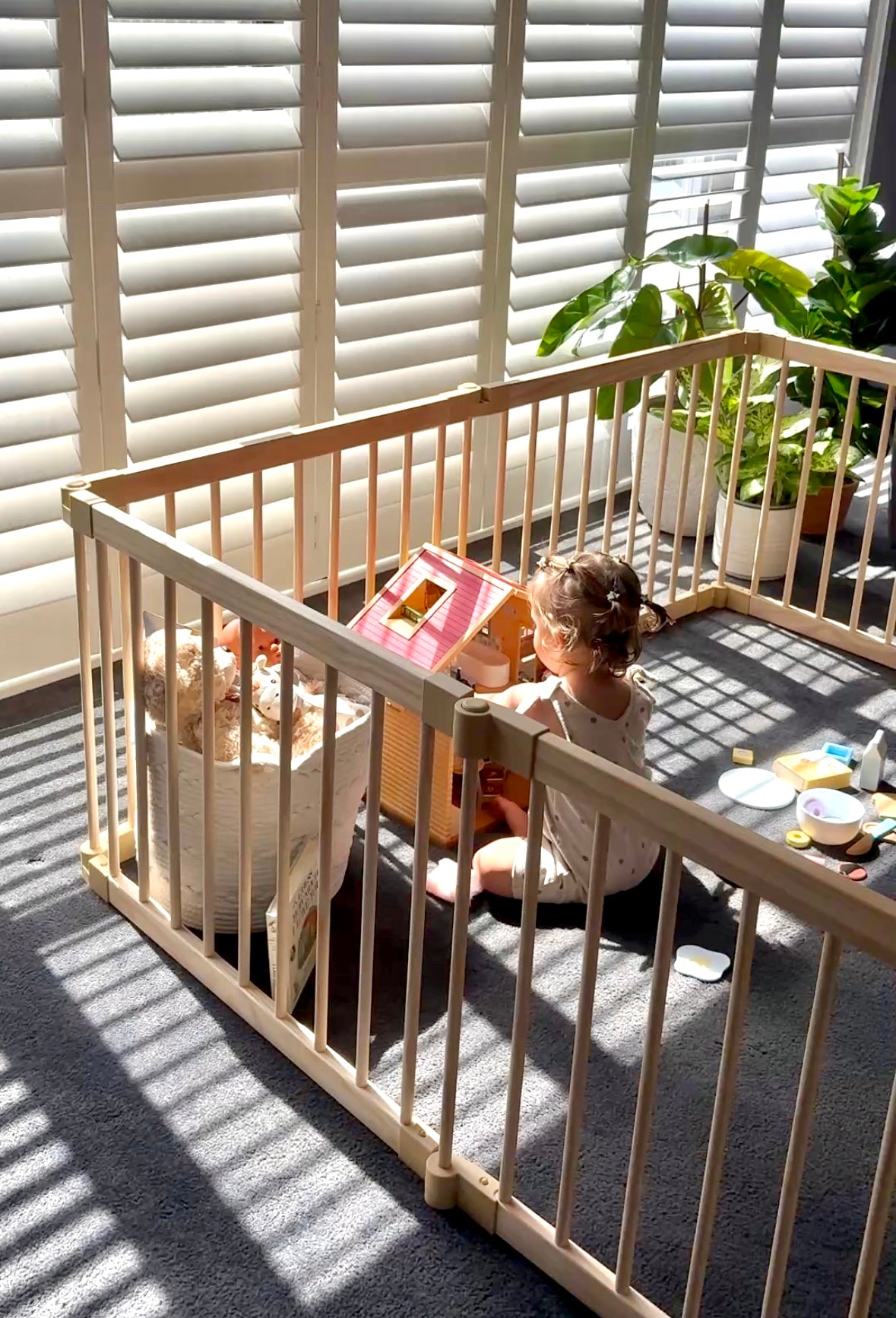 Child playing with toys behind a baby gate in a sunlit room.